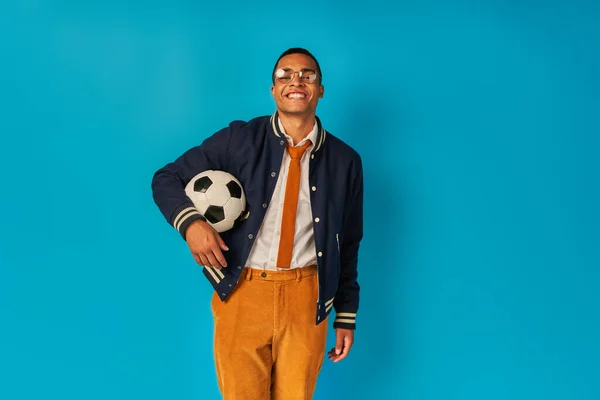 Estudiante afroamericano satisfecho en pantalones anaranjados, con pelota de fútbol, sonriendo a la cámara en azul - foto de stock