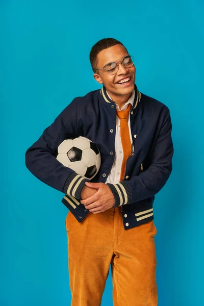 Estudiante afroamericano feliz en chaqueta elegante y pantalones anaranjados sosteniendo pelota de fútbol en azul - foto de stock