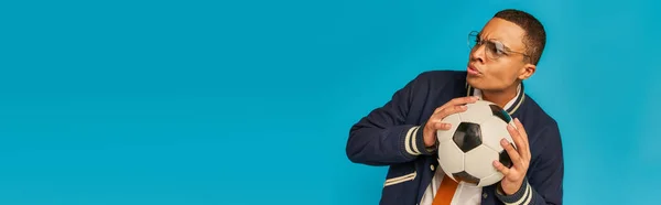 Serious african american student in eyeglasses looking away and holding soccer ball on blue, banner — Stock Photo