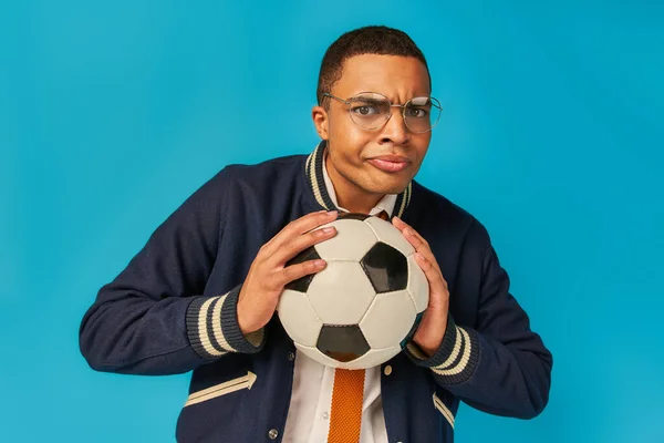 Serio africano americano studente in occhiali in possesso di palla da calcio e sbuffare guance su blu — Foto stock