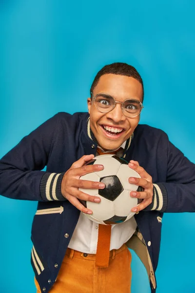 Overjoyed african american in jacket and eyeglasses holding soccer ball, smiling at camera on blue — Stock Photo