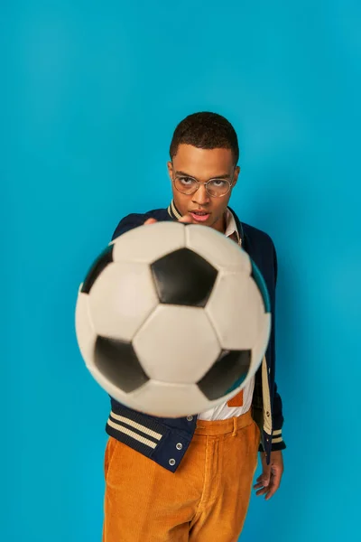 Confiant et sérieux afro-américain étudiant montrant ballon de football à la caméra sur bleu — Photo de stock
