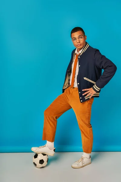 Estudante americano africano na moda na jaqueta, calças laranja pisando na bola de futebol no azul, mão no quadril — Fotografia de Stock