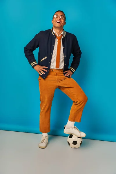 Excited and trendy african american student stepping on soccer ball and laughing on blue — Stock Photo