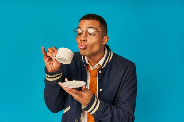 Африканский студент в стильной куртке и очках держит чашку кофе и блюдце на синем — стоковое фото