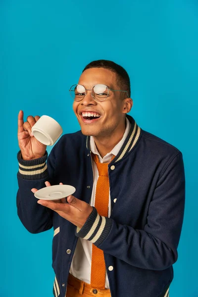 Allegro ed elegante studente afro-americano in occhiali da vista in possesso di tazza di caffè su sfondo blu — Foto stock