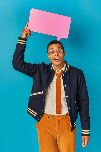 Усміхнений афроамериканський студент у модній куртці та окулярах, що тримає мовну бульбашку на синьому — стокове фото