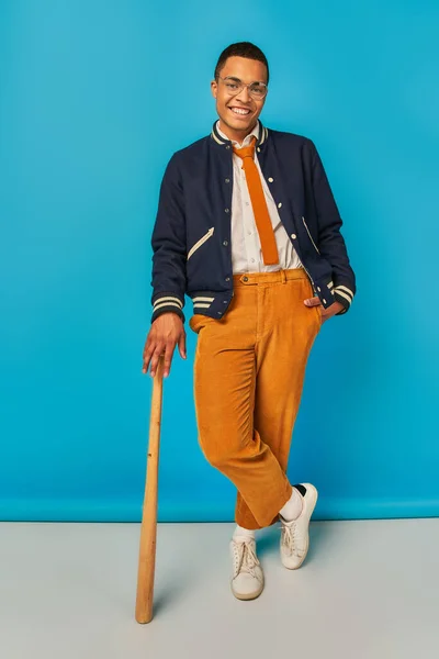 Estudiante afroamericano sonriente en chaqueta y pantalones naranja posando con bate de béisbol en azul - foto de stock
