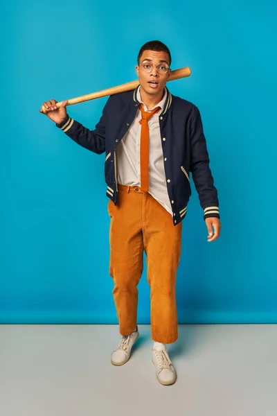 Étudiant afro-américain expressif en pantalon orange tenant batte de baseball, regardant caméra sur bleu — Photo de stock