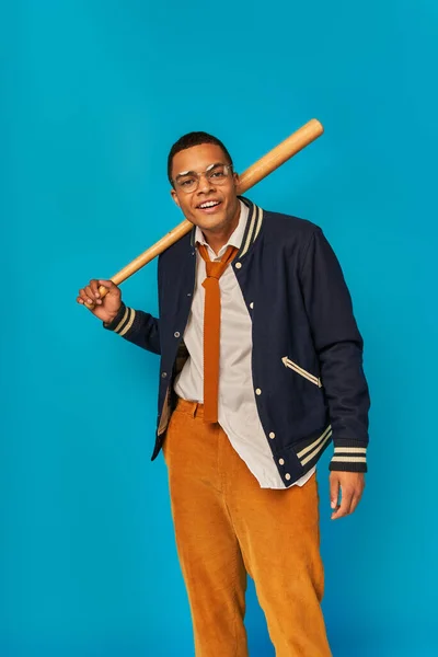 Estudiante afroamericano en pantalones naranja sosteniendo bate de béisbol y sonriendo a la cámara en azul - foto de stock