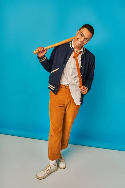Alegre y elegante estudiante afroamericano con bate de béisbol tirando de corbata en azul - foto de stock