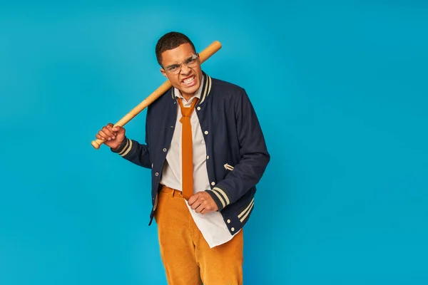 Étudiant afro-américain en colère dans des vêtements élégants tenant batte de baseball et grimaçant sur bleu — Photo de stock