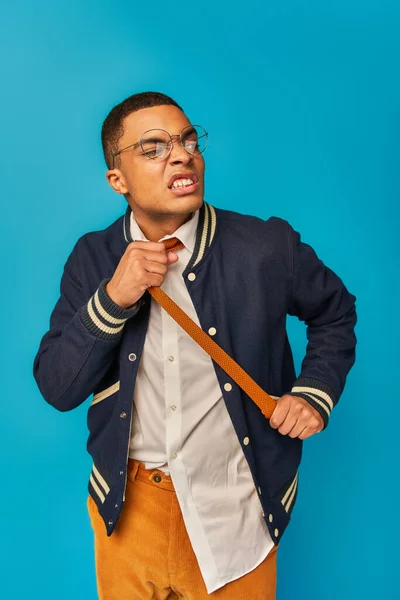 Displeased and angry african american student in eyeglasses pulling tie on blue — Stock Photo