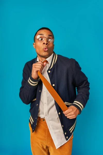 Estudiante afroamericano sorprendido haciendo pucheros labios, tirando de la corbata y mirando hacia otro lado en azul - foto de stock