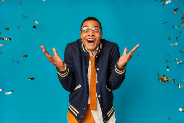Étudiant afro-américain joyeux et à la mode montrant wow geste lors de la fête sous confettis sur bleu — Photo de stock