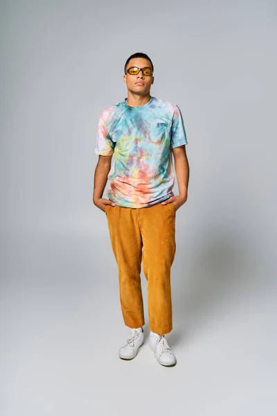 African american man in sunglasses, tie-dye t-shirt and orange pants with hands in pockets on grey — Stock Photo