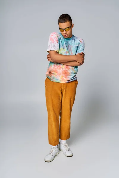 Homme afro-américain à la mode avec les mains pliées sur le gris, t-shirt tie-dye, pantalon orange, lunettes de soleil — Photo de stock