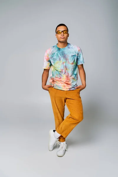 Homme afro-américain à la mode avec les mains dans les poches sur gris, pantalon orange, lunettes de soleil, t-shirt tie-dye — Photo de stock