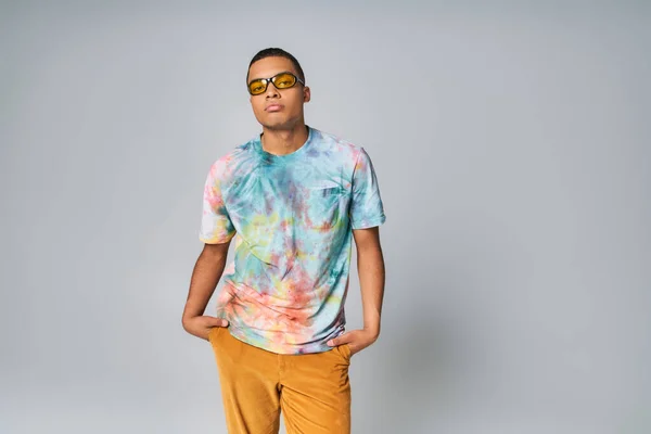 Young african american man in sunglasses, tie-dye t-shirt, orange pants on grey — Stock Photo