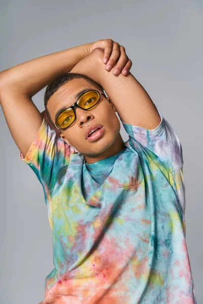 Homme afro-américain avec les mains au-dessus de la tête regardant caméra sur gris, lunettes de soleil, t-shirt tie-dye — Photo de stock