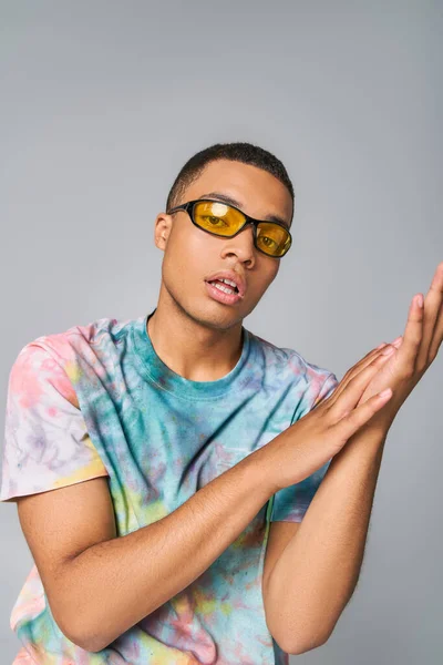 Jeune homme afro-américain élégant en t-shirt tie-dye et lunettes de soleil regardant caméra sur gris — Photo de stock