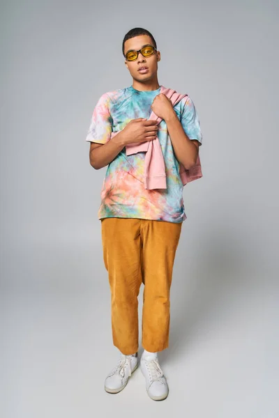 Homme afro-américain à la mode, lunettes de soleil, t-shirt tie-dye, pantalon orange sur gris, pleine longueur — Photo de stock