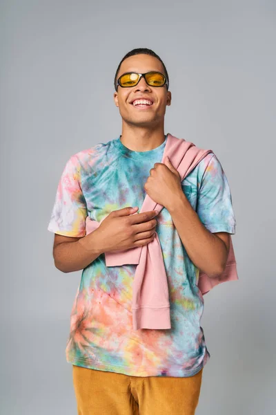Happy african american man in sunglasses and tie-dye t-shirt looking at camera on grey — Stock Photo
