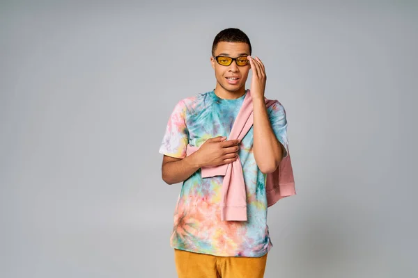 Trendy african american man in orange pants and tie-dye t-shirt adjusting sunglasses on grey — Stock Photo