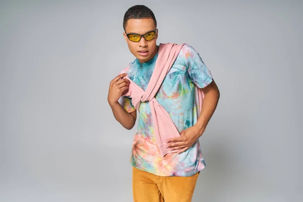 African american man in sunglasses and tie-dye t-shirt posing with hand on hip on grey — Stock Photo