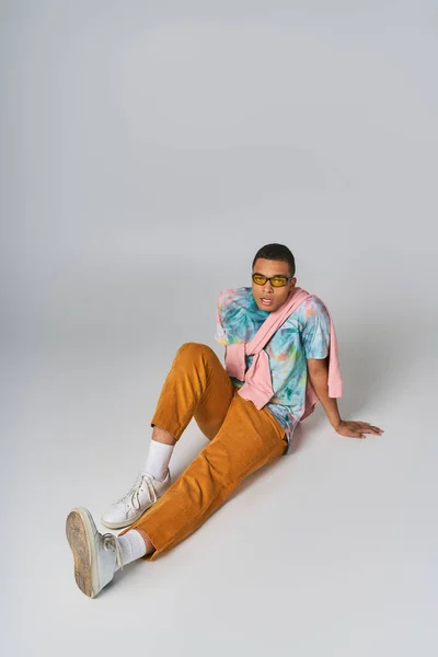 Trendy african american man sitting on grey, sunglasses, orange pants, tie-dye t-shirt — Stock Photo