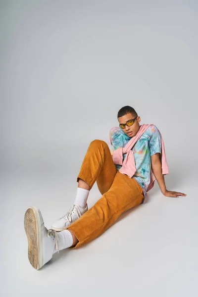 Homme afro-américain en t-shirt tie-dye, pantalon orange et lunettes de soleil assis sur gris — Photo de stock