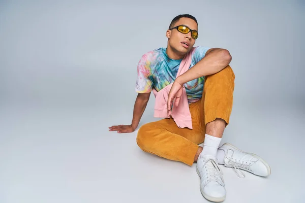 Confident african american man in orange pants, tie-dye t-shirt and sunglasses sitting on grey — Stock Photo