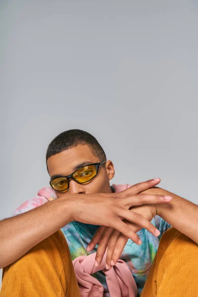 Jeune homme afro-américain en lunettes de soleil à la mode et t-shirt tie-dye regardant la caméra sur gris — Photo de stock
