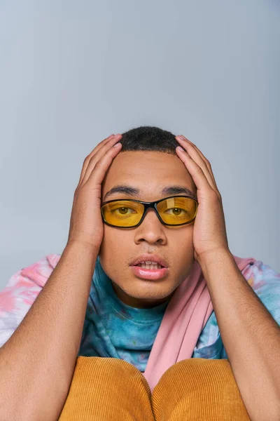 Homme afro-américain à la mode en lunettes de soleil, t-shirt tie-dye toucher la tête, en regardant la caméra sur gris — Photo de stock