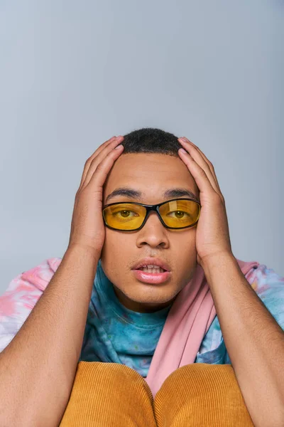 Trendy african american man in sunglasses, tie-dye t-shirt touching head, looking at camera on grey — Stock Photo