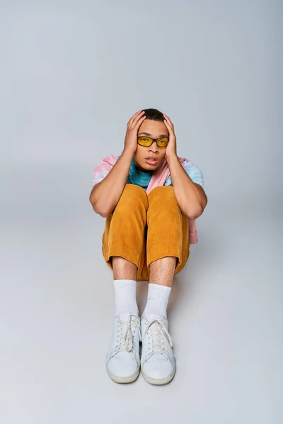 African american man in trendy clothes touching head and looking at camera while sitting on grey — Stock Photo