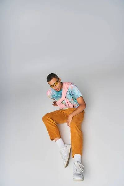 Afro-américain en pantalon orange, t-shirt tie-dye, lunettes de soleil assises sur gris, vue grand angle — Photo de stock