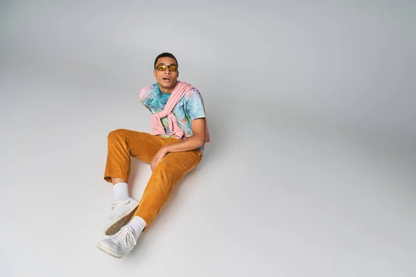 African american man in orange pants, tie-dye t-shirt, sunglasses, looking at camera on grey — Stock Photo
