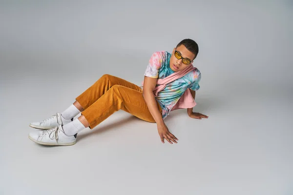 Uomo afroamericano, pantaloni arancioni, t-shirt tinta unita, occhiali da sole seduti sul grigio, guardando la macchina fotografica — Foto stock