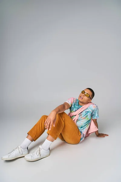 Chico afroamericano, pantalones anaranjados, corbata-tinte camiseta, gafas de sol sentado en gris, mirando hacia otro lado - foto de stock