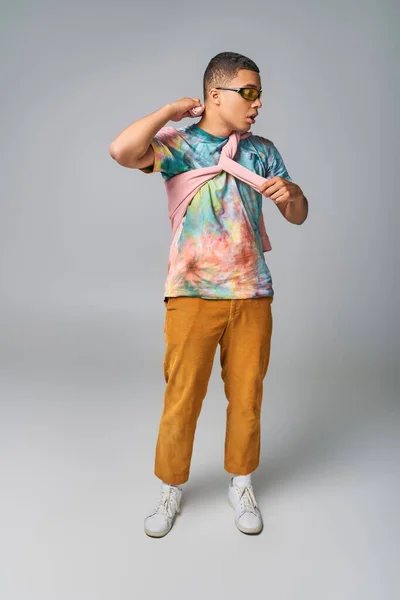 African american man, orange pants, tie-dye t-shirt, sunglasses posing on grey, looking away — Stock Photo