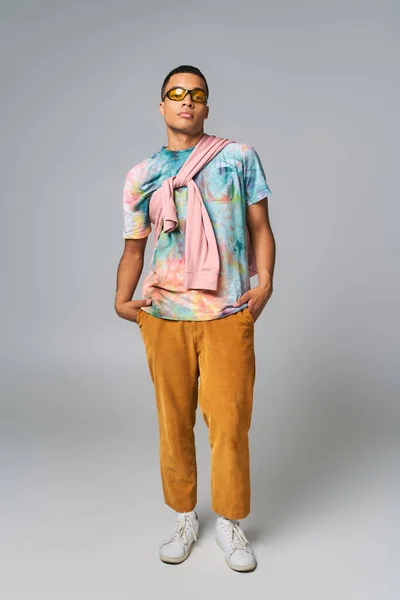 Homme afro-américain confiant, pantalon orange, lunettes de soleil, t-shirt tie-dye, les mains dans la poche, sur gris — Photo de stock