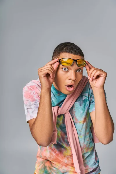Amazed african american man in tie-dye shirt holding sunglasses and looking at camera on grey — Stock Photo