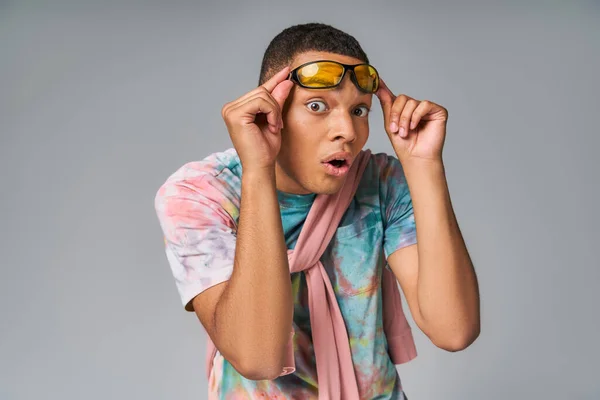 Surpreso afro-americano cara em tie-dye camisa segurando óculos de sol e olhando para a câmera em cinza — Fotografia de Stock