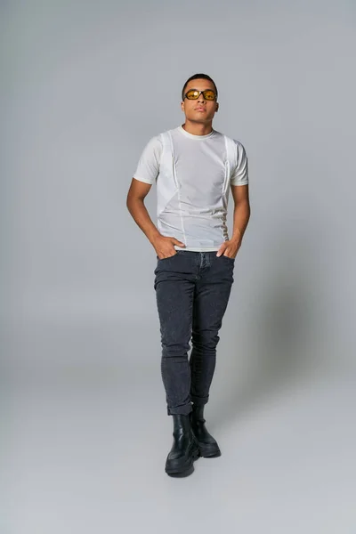 Homme afro-américain à la mode en lunettes de soleil, t-shirt, jeans regardant la caméra sur gris — Photo de stock