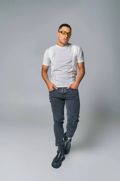 Elegante uomo afroamericano in occhiali da sole, t-shirt, jeans guardando la fotocamera sul grigio — Foto stock