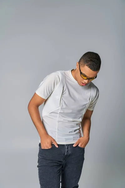 Joven hombre afroamericano en gafas de sol de moda, camiseta, jeans, con las manos en los bolsillos en gris - foto de stock