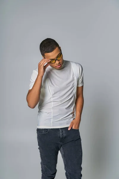 Trendy african american man in t-shirt and jeans, with hand in pocket, adjusting sunglasses on grey — Stock Photo