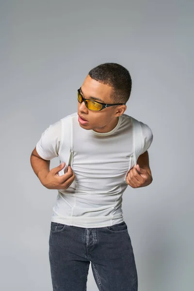 Youthful african american man in stylish t-shirt and sunglasses looking away on grey — Stock Photo