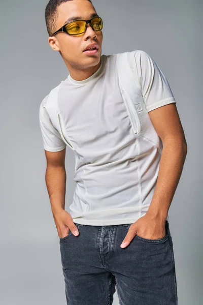 Trendy african american man in stylish t-shirt and sunglasses looking away on grey, hands in pockets — Stock Photo